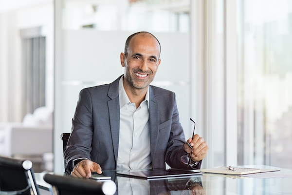 A middle aged in a business suit holding a pair of glasses smiles at the camera