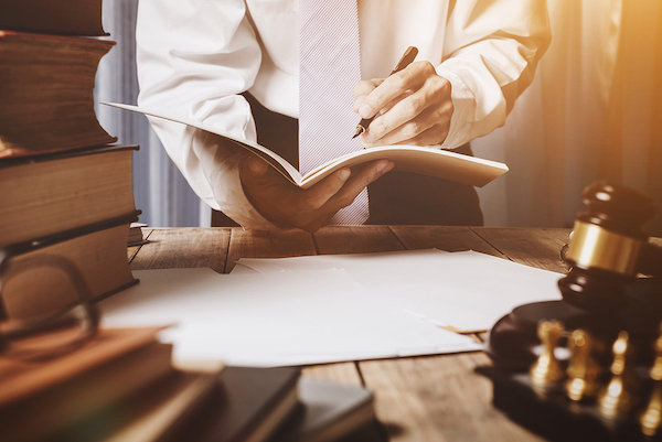 A man in a suit writes in a notebook standing near a desk containing law books and a gavel